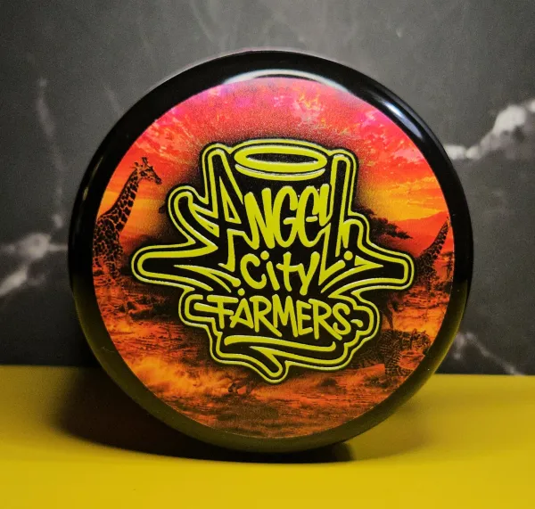 Top view of an Angel City Farmers Giraffe Puzzy jar lid featuring neon yellow graffiti branding over a safari sunset background.