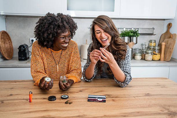 Two women sitting at a wooden kitchen counter laughing while inspecting a pre-roll joint and dispensary stash jar of Giraffe Puzzy strain.
